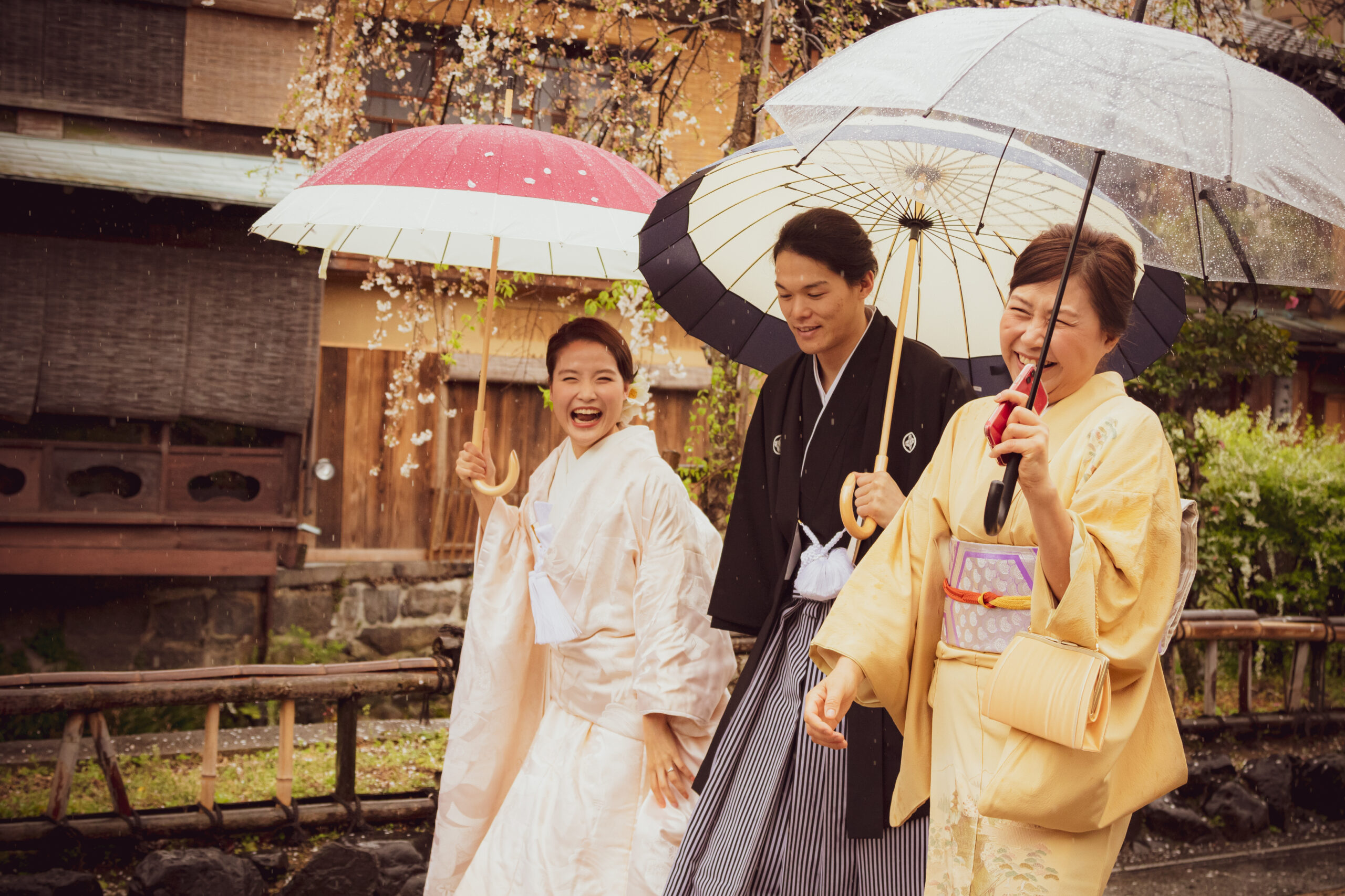 雨の中京都の街並みで和装を着て和傘をさす新郎新婦と和装の新郎の母の3人でお散歩している