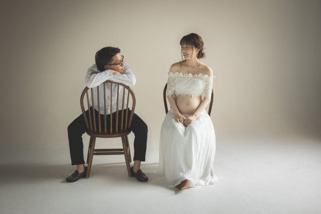白いオフショルダーの衣装でお腹を見せた妻と見つめ合うパートナーのマタニティフォト