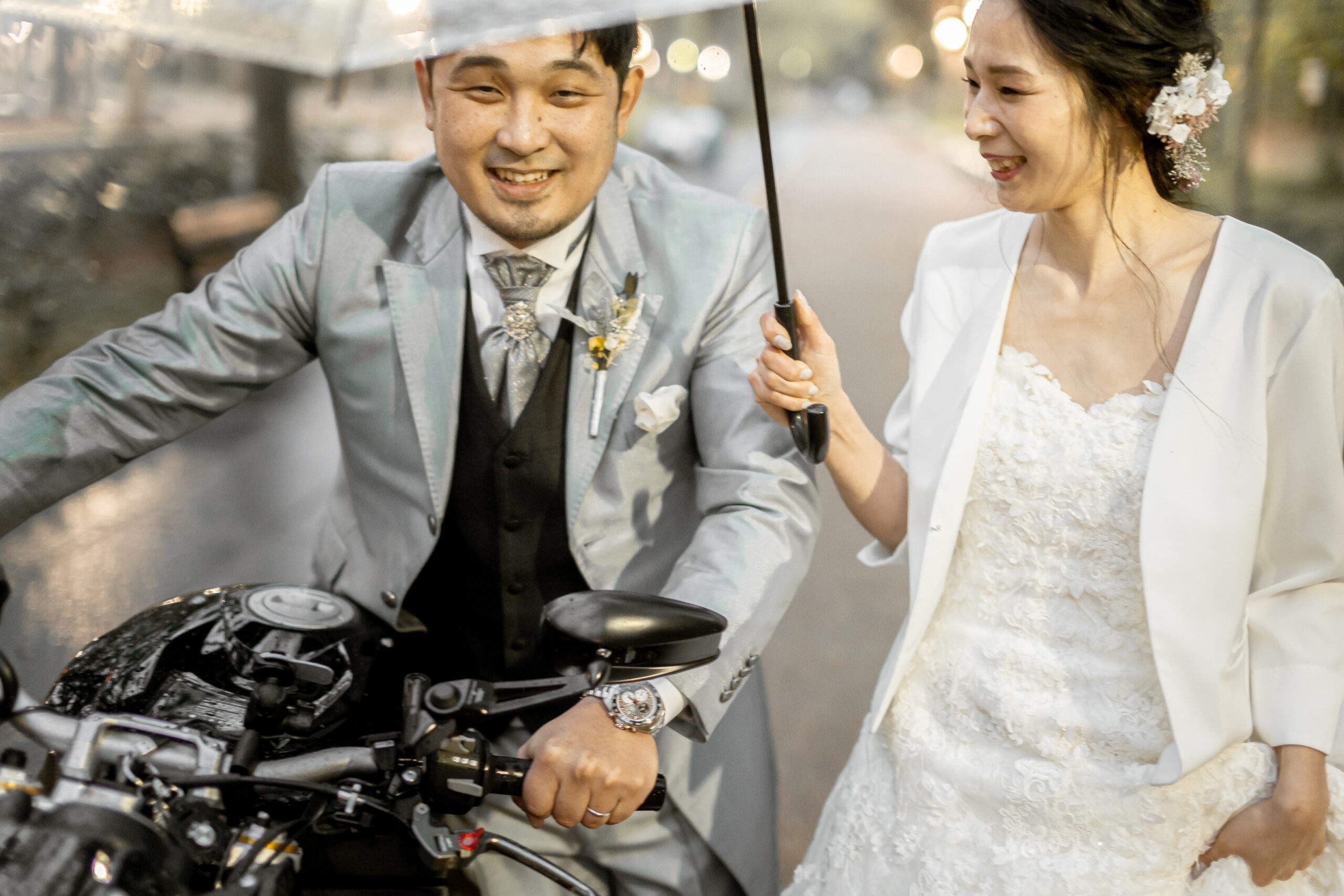 バイクを手押しするシルバータキシードの新郎とウェディングドレスの新婦が相合い傘