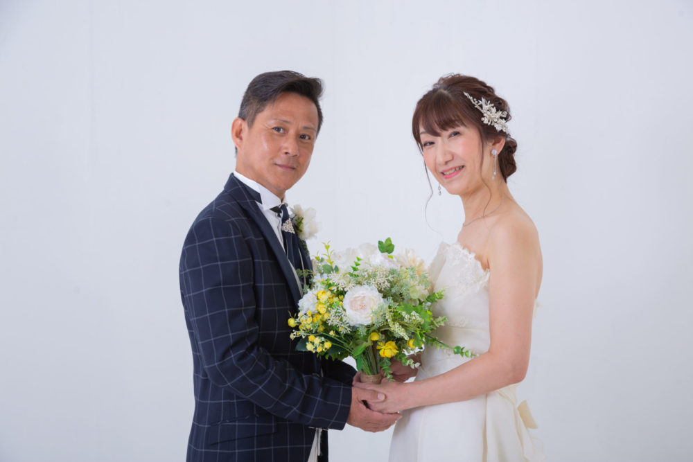 ウェディングドレスとタキシードを着た大人夫婦が2人でブーケをもちこちらに微笑みかけている