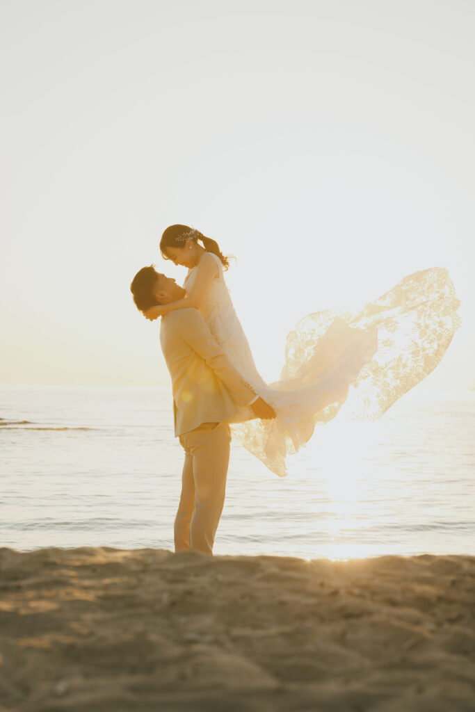 大阪フォトウェディング海ロケで日の入りの太陽が水面に反射する中新郎が新婦を抱っこしドレスのトレーンがなびくシーン