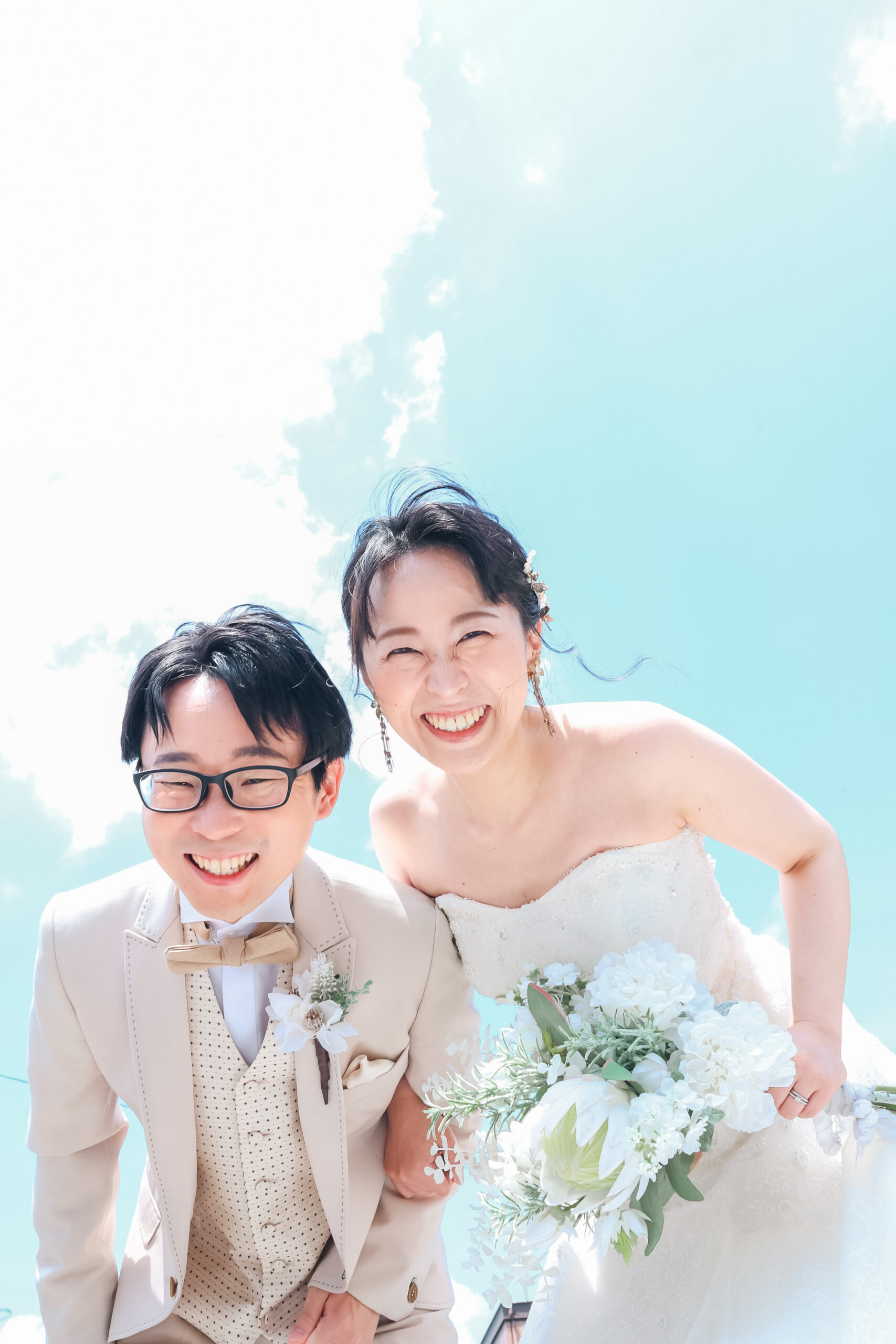 青空をバックにカメラをのぞき込むように笑顔の新郎新婦